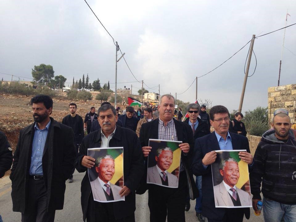 Protesters, among them Hadash chairman MK Muhammad Barakeh, holding a singe with Nelson Mandela portrait during a demonstration held in Nabi Saleh on December 7, 2013 to commemorate the killing of Mustafa Tamimi and Rushdi Tamimi by Israeli military occupation forces (Photo: Al Ittihad)