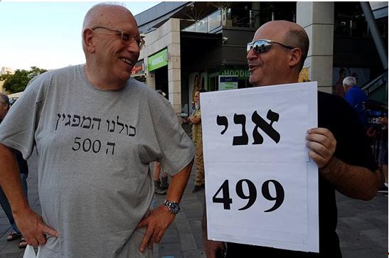 Two of the some 2,000 protestors who turned out on Saturday evening in Petah Tikva, make light of the supposed limit of 500 set for the event: "I’m demonstrator number 499" declares one of them in the sign he’s holding, and to the left the grey t-shirt reads: “All of us are demonstrator number 500.”