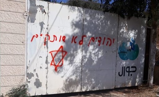 A threat spray-painted in red "Jews don't remain silent" defaces the entrance gate to a building in the occupied West Bank town of Yatma, 15km south of Nablus, August 13, 2019. The building serves as offices to Jawwal, identified by its logo to the right, a major Palestinian-owned cellular communications company.