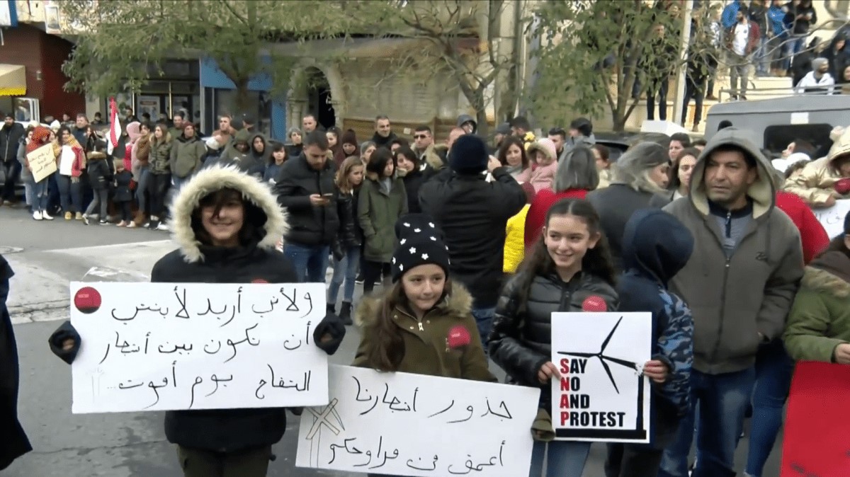 A demonstration held in Majdal Shams against Israeli plans for a commercial wind farm that would see at least 25 turbines built in the occupied Syrian territory. The placard in Arabic on the right reads: "The roots of our trees are stronger than your wind turbines"; the sign on the left says: "Because I want my daughter to be among the apple trees when I die."