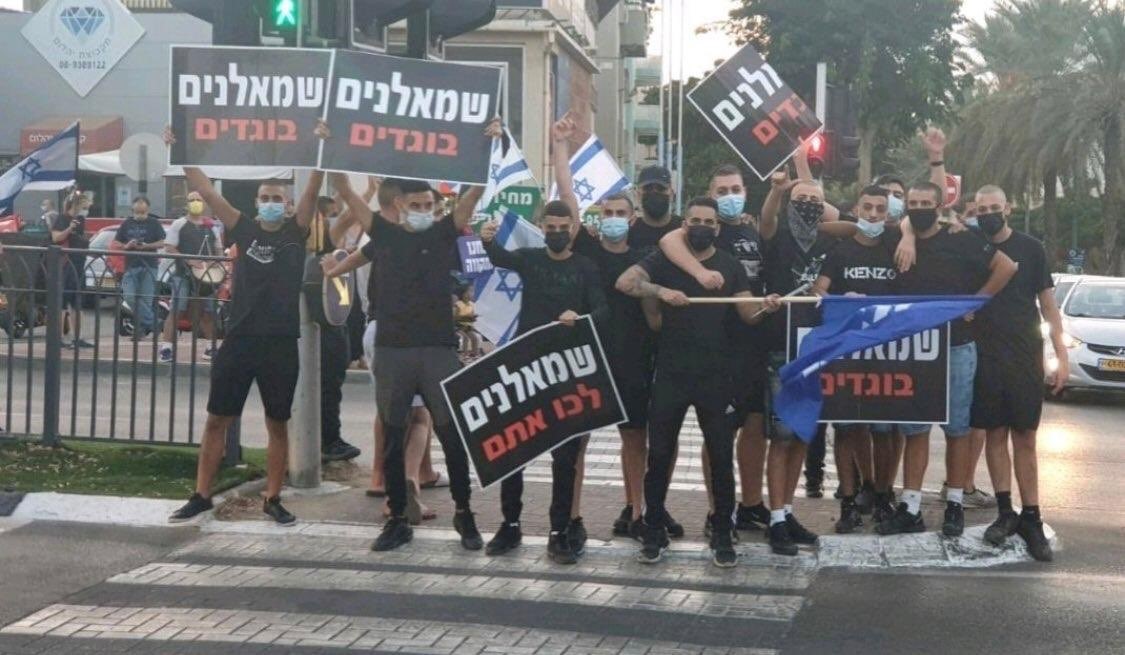 Members of the La Familia gang brandish a blue Likud flag and placards reading "Leftists are traitors" and "Leftists, go with them" [i.e., leave with the Arabs] opposite an anti-Netanyahu demonstration in Rabin Square, Tel Aviv, October 17, 2020.