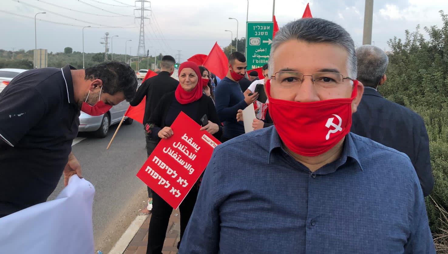 MK Youssef Jabareen (Hadash-Joint List) during a protest organized by the front in the Galilee; the Hadash placard by a woman protester in the mid-ground reads in Arabic "Down with the occupation and exploitation"; in Hebrew it reads "No to annexation, no to discrimination."
