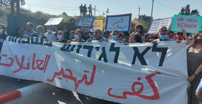 Activists demonstrate alongside local residents against the planned evictions of Palestinian refugee families living since 1956 in the occupied East Jerusalem neighborhood of Sheikh Jarrah, Friday, April 16, 2021. The large banner reads in Hebrew and Arabic “No to the displacement of families.” An Arabic placard held aloft to the right of center says “Displacement is a war crime.”