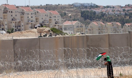 A woman places a Palestinian flag on the barbed wire fence blocking access to the concrete Separation Wall beyond which is Israel's illegal settlement in Modi’in Illit.