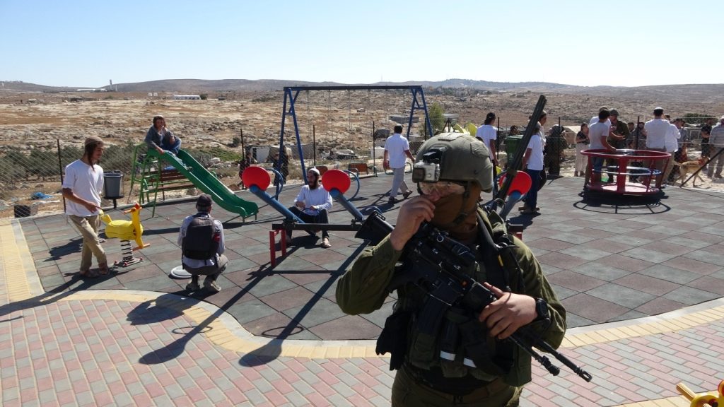 An occupation soldier stands guard over Israeli settlers after they invaded a playground in the Palestinian hamlet of Susya in the West Bank's South Hebron Hills, chasing off the village's children, Saturday, November 6, 2021.